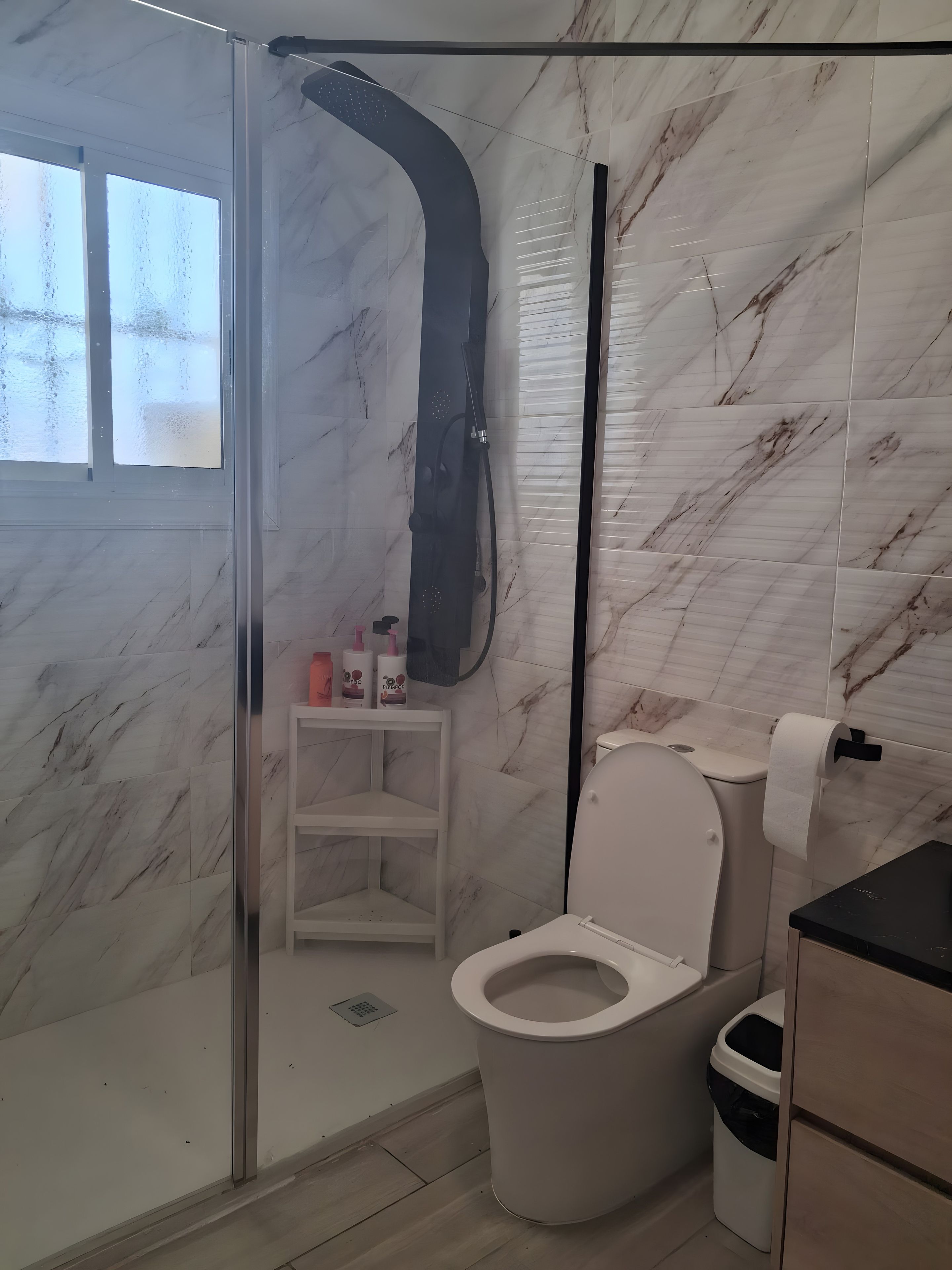 A bathroom with a shower enclosure, toilet, and a shelf with toiletries.