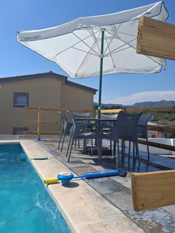 A poolside area with a table and chairs, a white umbrella, and a view of mountains.