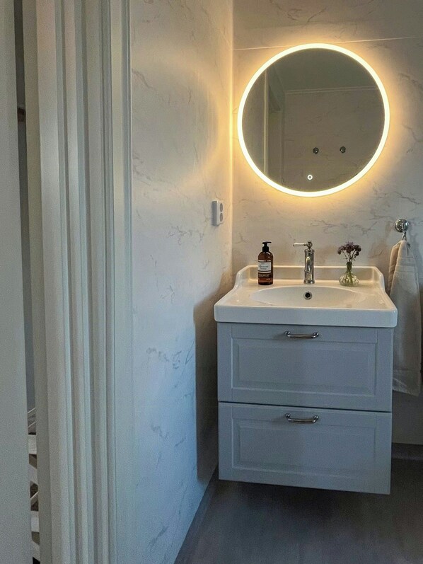 A bathroom with a white vanity, a round mirror with a light, a faucet, a bottle of soap, and a towel hanging on the wall.