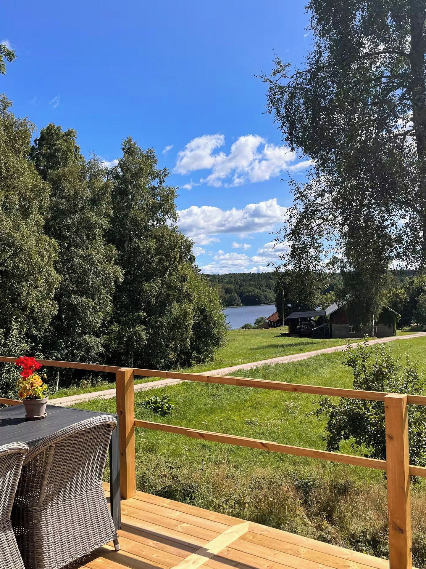 A wooden deck with a wicker chair and a table, overlooking a lake and a cabin.