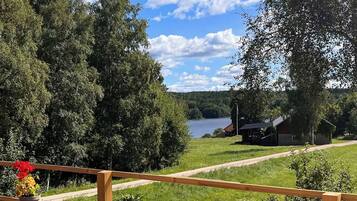 A wooden deck with a wicker chair and a table, overlooking a lake and a cabin.