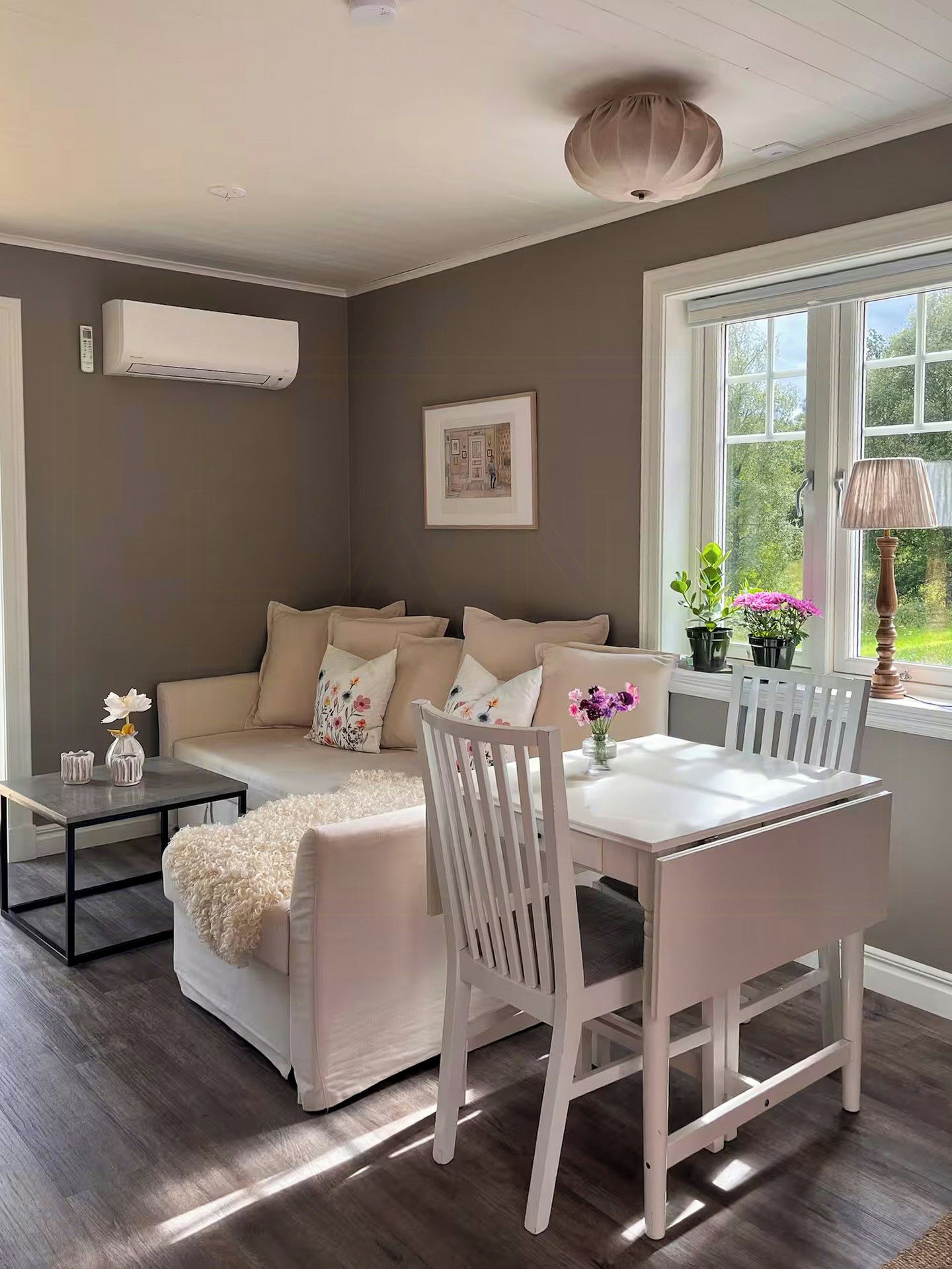 A cozy living room with a white sofa, a dining table, and chairs. There is a window with a view of greenery outside.