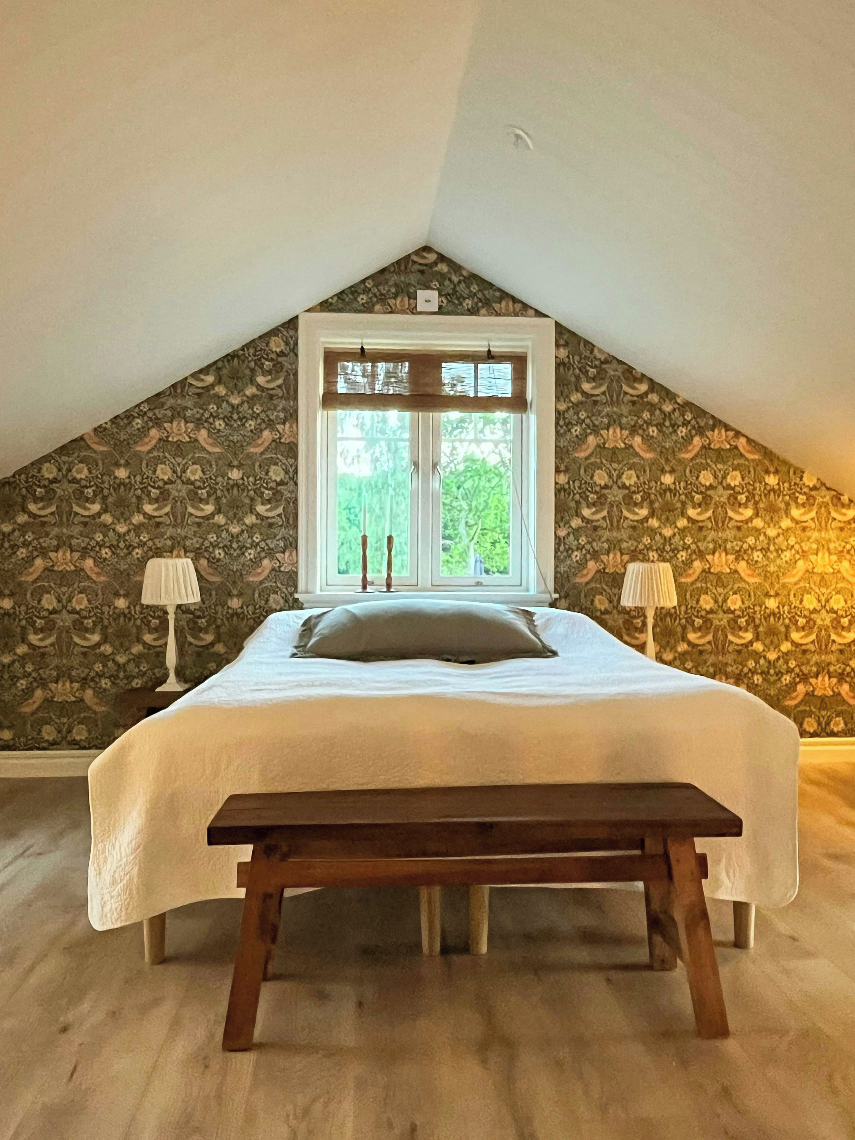 A bedroom with a sloped ceiling, a bed with a white bedspread, two bedside lamps, and a wooden bench.