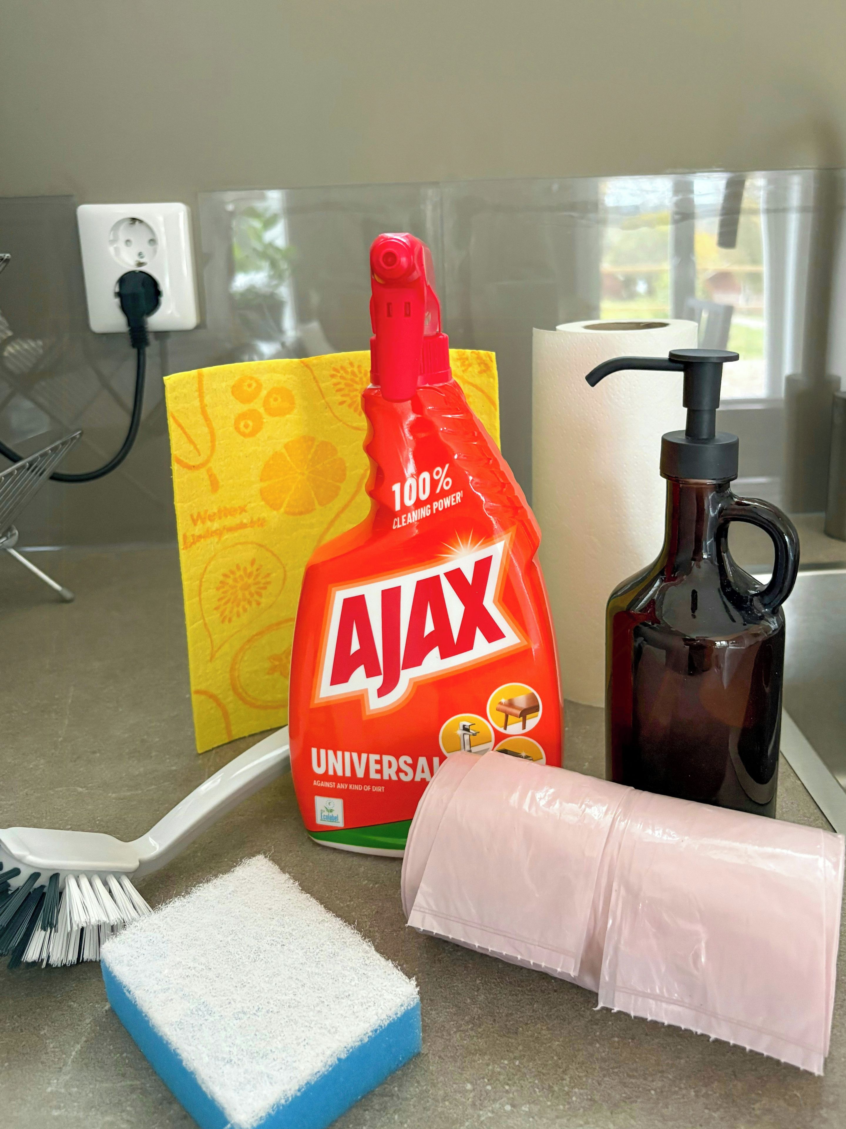 Una encimera de cocina con productos de limpieza, una botella de Ajax Universal, una botella negra, un paño rosa y un cepillo blanco.
