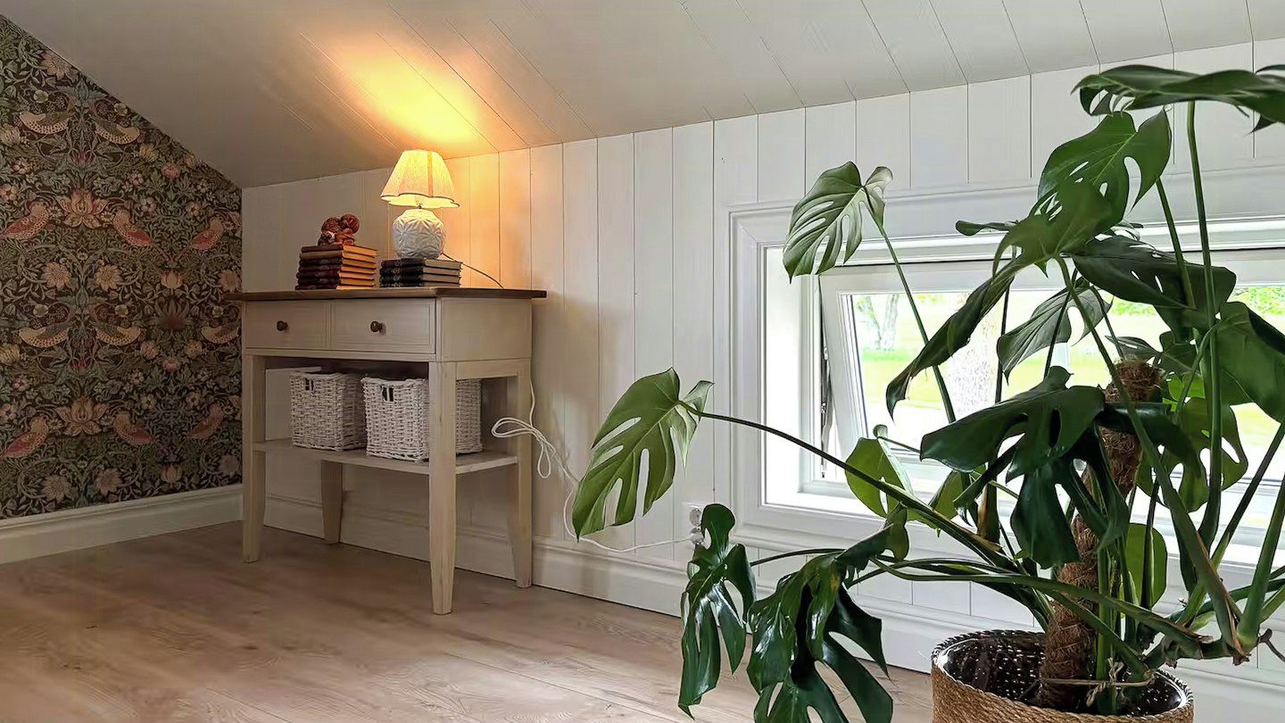 A room with a wooden floor, a white wall with floral wallpaper, a bedside table with a lamp, books, and baskets, and a large plant in a woven pot.