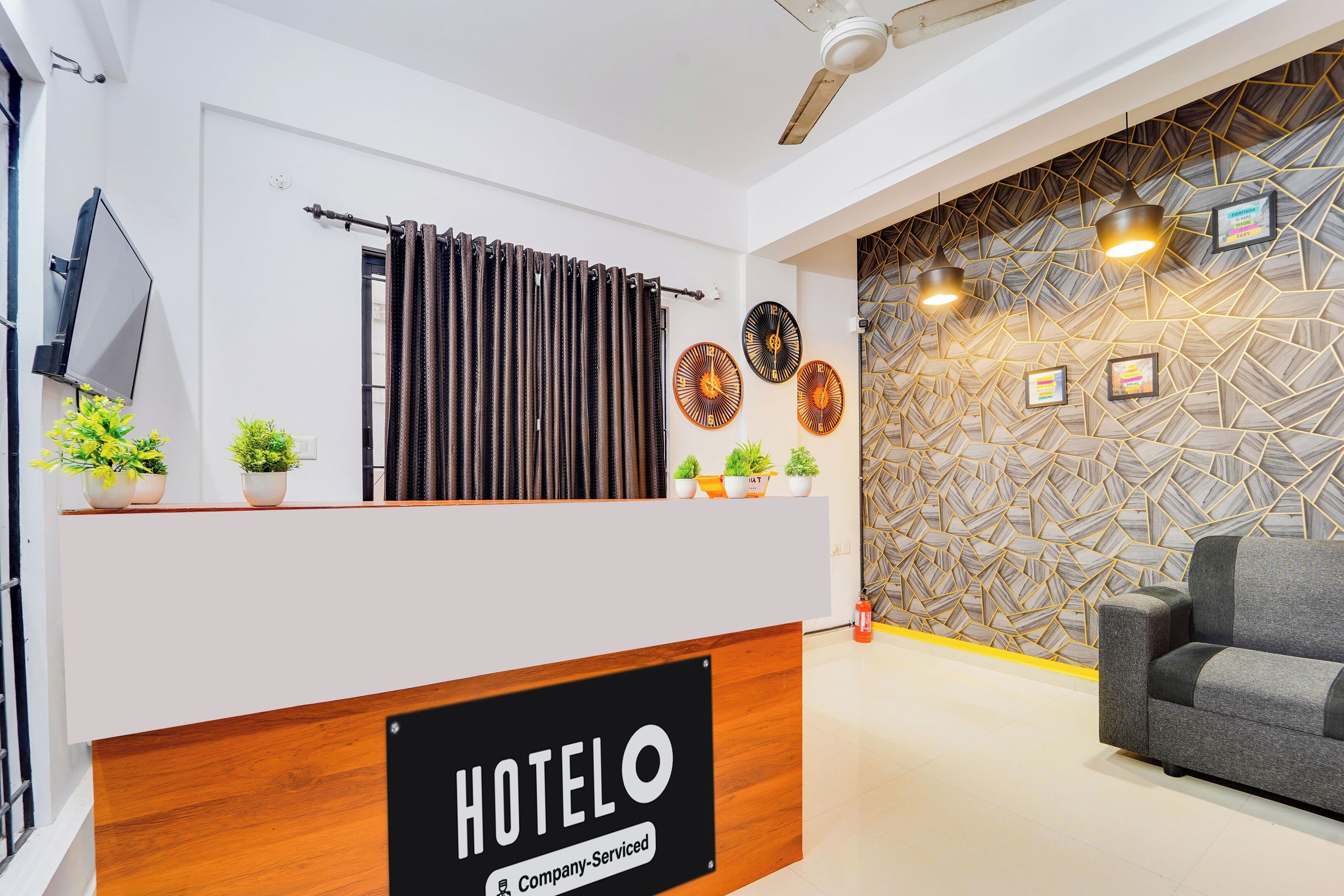 Un comptoir de réception d’hôtel avec une enseigne indiquant « HOTELO » et une étiquette « Service d’entreprise ». On y trouve trois horloges murales, un téléviseur à écran plat et un mur décoré derrière le comptoir.