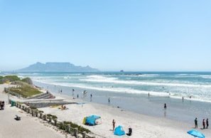 A beach with a clear sky, a sandy shore, and a few tents and umbrellas. - Serene 1-bedroom apartment in vibrant Big Bay neighborhood (Cape Town)