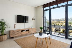 A modern living room with a flat-screen TV, a wooden entertainment unit, a round table with a potted plant, and large windows offering a view of the city. - Serene 1-bedroom apartment in vibrant Big Bay neighborhood (Cape Town)