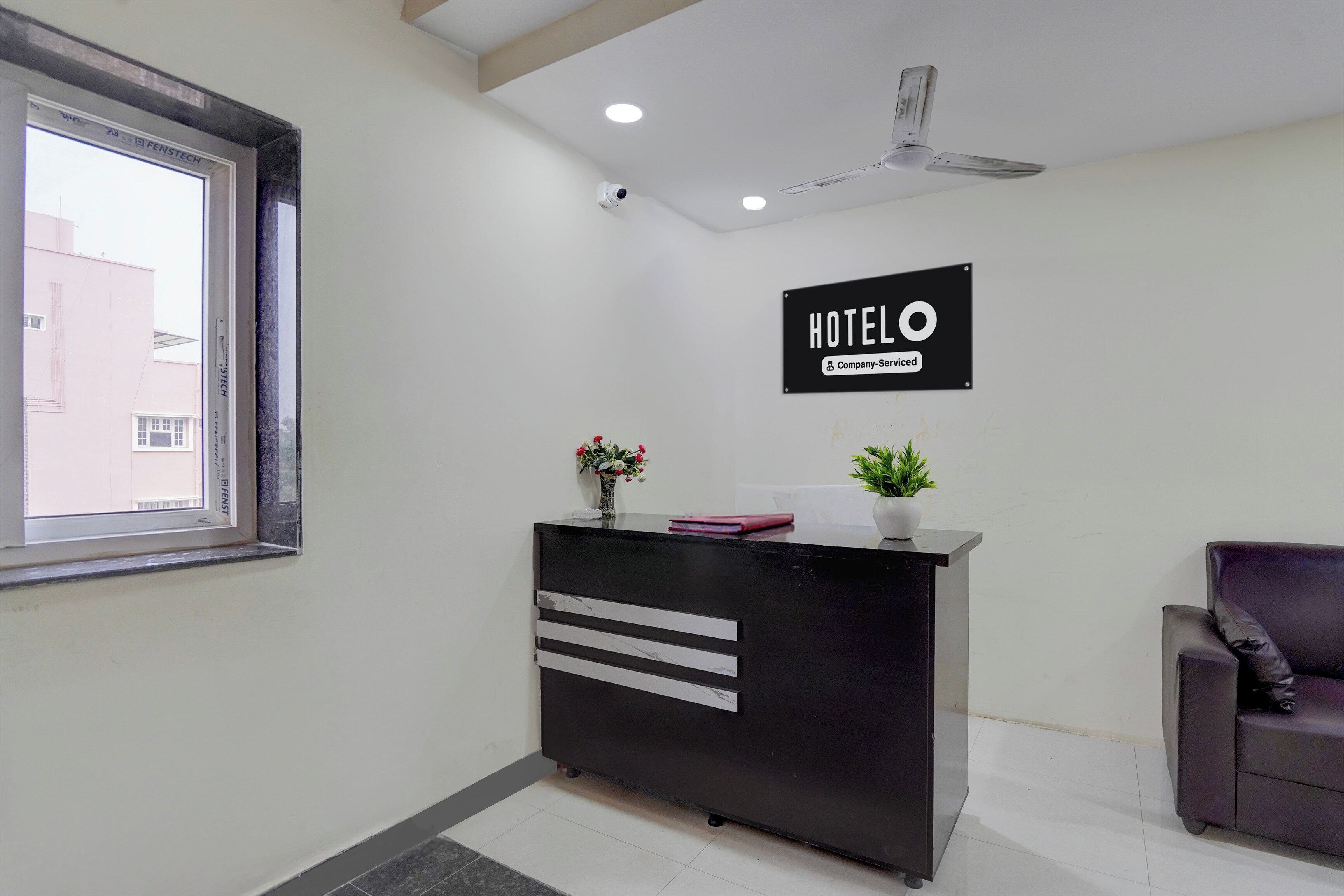 A hotel reception desk with a sign indicating it is company-serviced, a ceiling fan, and a window with a view of buildings outside.