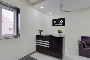 A hotel reception desk with a sign indicating it is company-serviced, a ceiling fan, and a window with a view of buildings outside.