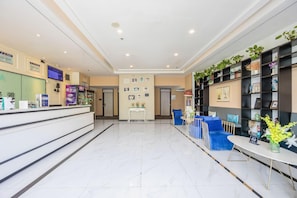 Zona de recepción de un hotel con suelo de mármol, mostrador de recepción, área de descanso con sillas azules y una estantería llena de diversos objetos.