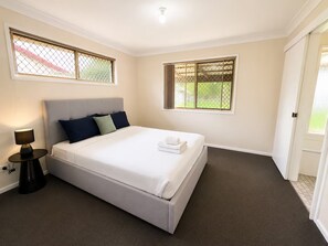 A hotel room with a bed, a bedside table with a lamp, and two windows with metal grilles. - entire 3BR Home | 3 Queen Beds, Garage & Fenced (Crestmead)