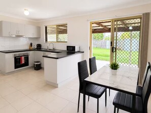 A modern kitchen with a dining area, featuring a large island, a built-in oven, and a view through a sliding glass door. - entire 3BR Home | 3 Queen Beds, Garage & Fenced (Crestmead)