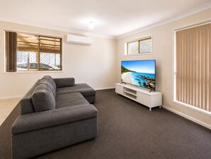 Ein modernes Wohnzimmer mit einem Ecksofa, einem Flachbildfernseher auf einem weißen Standfuß und einem großen Fenster mit Jalousien.