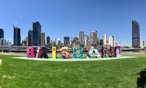 Ein grünes Feld im Vordergrund mit großen, farbenfrohen Buchstaben, die 'BRISBANE' bilden, mit einer Stadtansicht im Hintergrund.