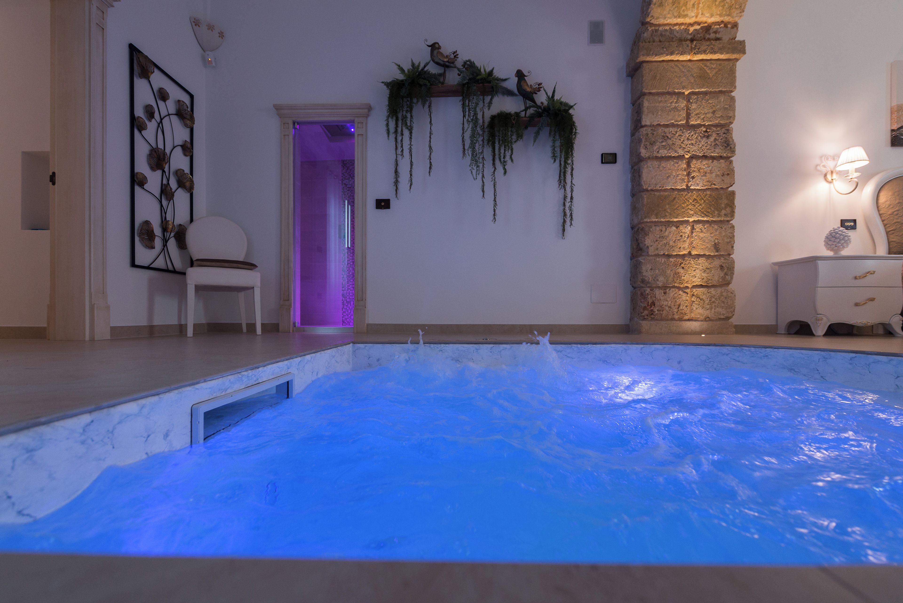 Une piscine dont l’intérieur est illuminé de lumière bleue, une chaise blanche, une porte donnant sur une pièce éclairée de lumière violette, et des œuvres d’art décoratives sur les murs.