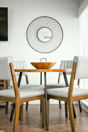 A dining area with a round table, four chairs with wooden frames and grey upholstery, and a wall-mounted circular mirror. - Roomy and Modern Apartment | Near AT&T Stadium (Arlington)