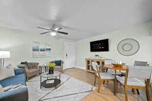 A living room with a ceiling fan, a flat-screen TV mounted on the wall, and a dining area with a wooden table and chairs. - Roomy and Modern Apartment | Near AT&T Stadium (Arlington)