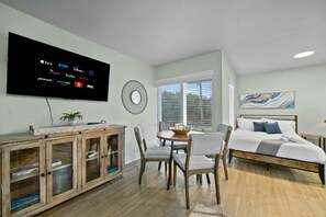 Une chambre moderne avec un lit, un coin repas avec des chaises, un téléviseur diffusant des services de streaming et une fenêtre avec des stores.