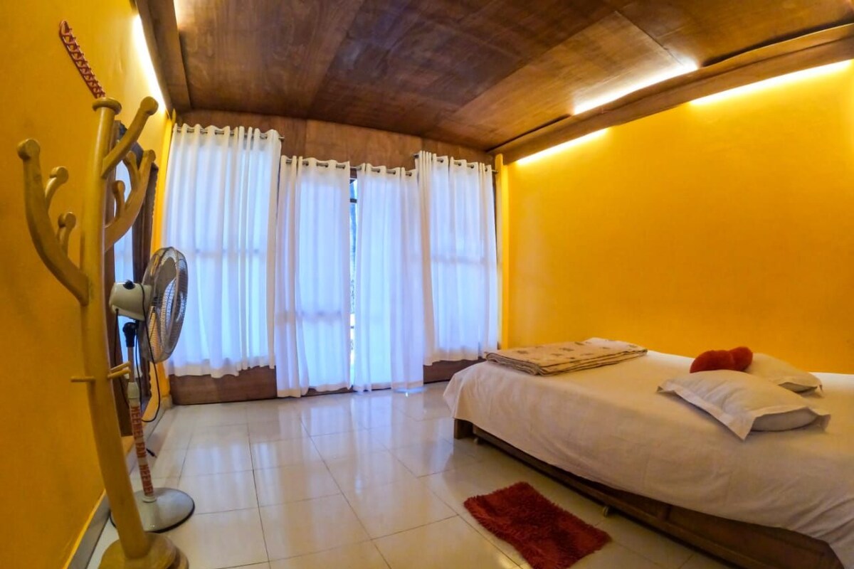 Quarto com cama, teto de madeira, ventilador e janela com cortinas.