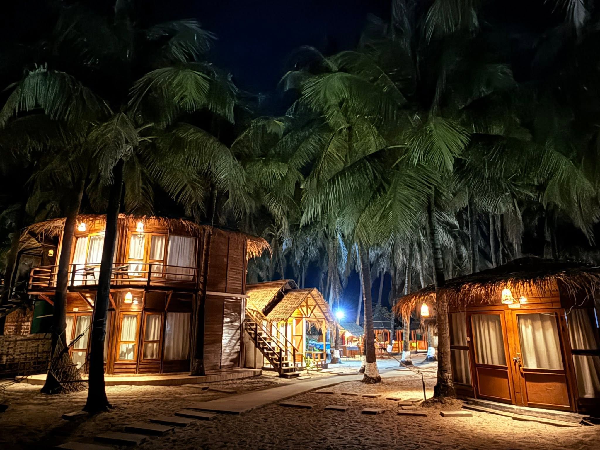 A beachfront resort with thatched-roof cabins, palm trees, and a sandy walkway.