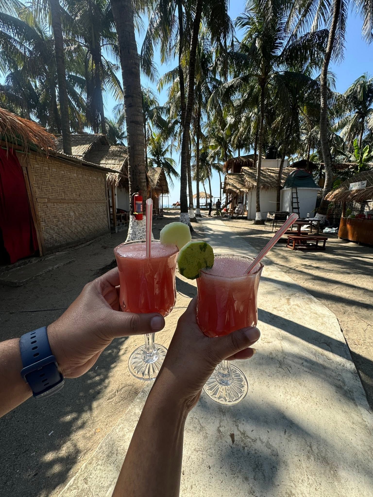 Two hands holding glasses of a red beverage with a lime garnish, set against a backdrop of palm trees and a beach setting.