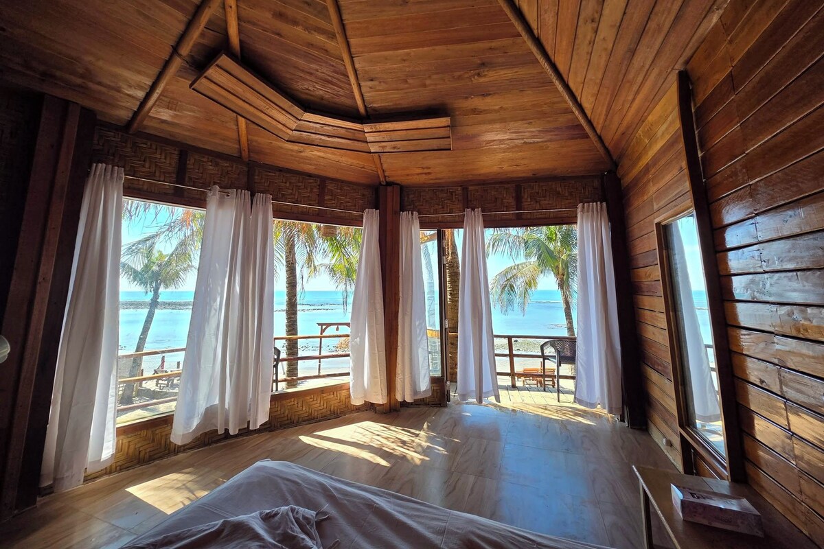 Uma cabana de madeira com cama, janelas amplas e vista para a praia.