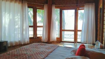 A room with a large bed, two windows with curtains, a view of palm trees, and a wooden ceiling.