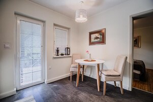 Una habitación con suelo de madera oscura, una mesa blanca con dos sillas, una lámpara colgante, una ventana con persianas y un cuadro enmarcado en la pared.