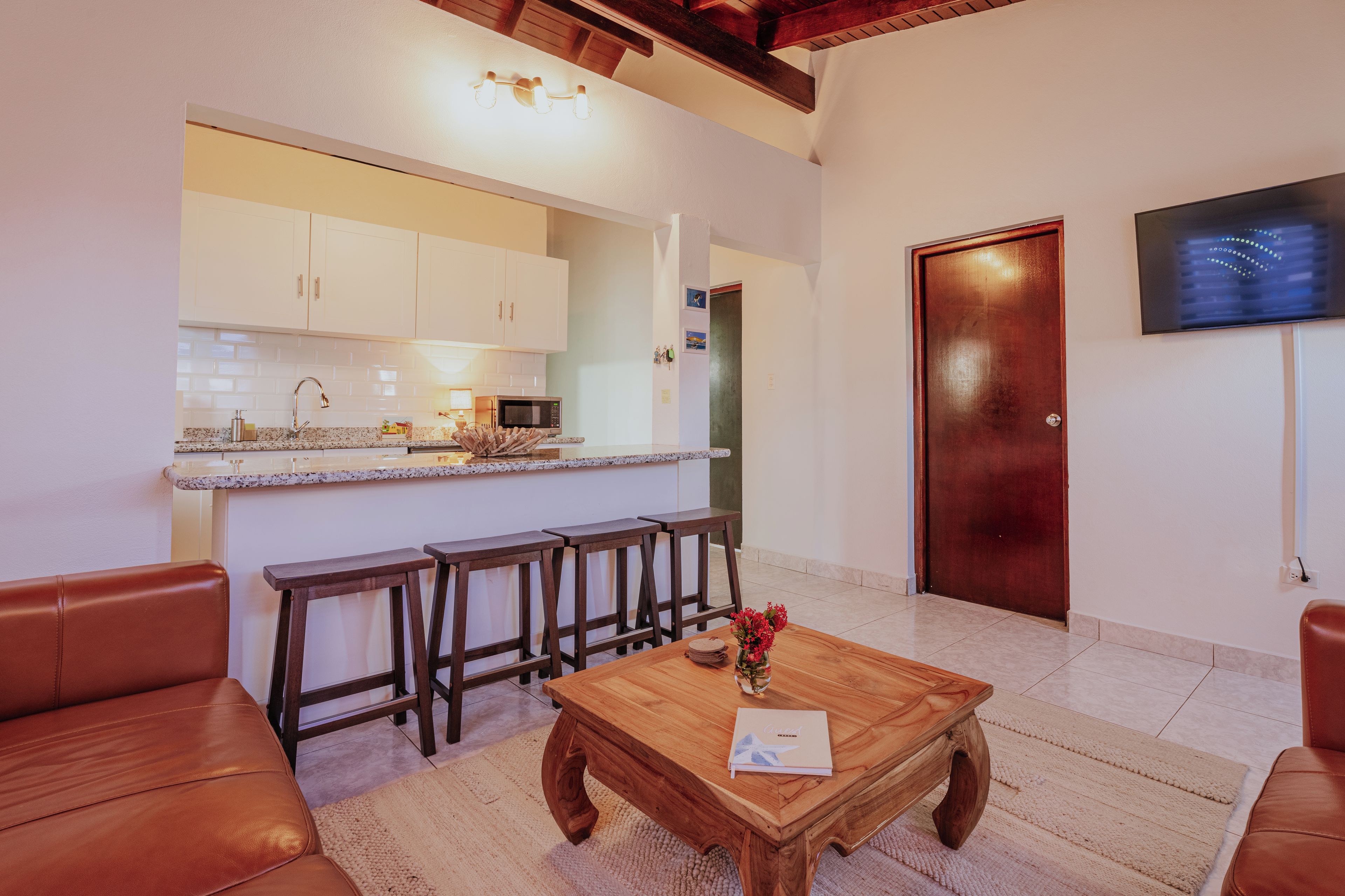 Ein modernes Wohnzimmer mit Couchtisch aus Holz, Ledersofa und Küchenzeile mit weißen Schränken und einer Mikrowelle.