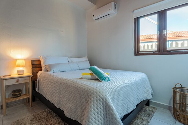 A bedroom with a bed, a bedside table with a lamp, a window, and an air conditioning unit.