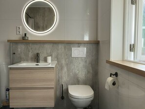 A modern bathroom with a round mirror, a sink, a toilet, and a window. - Holiday Home in Algana With Swimming Pool (Årjäng)