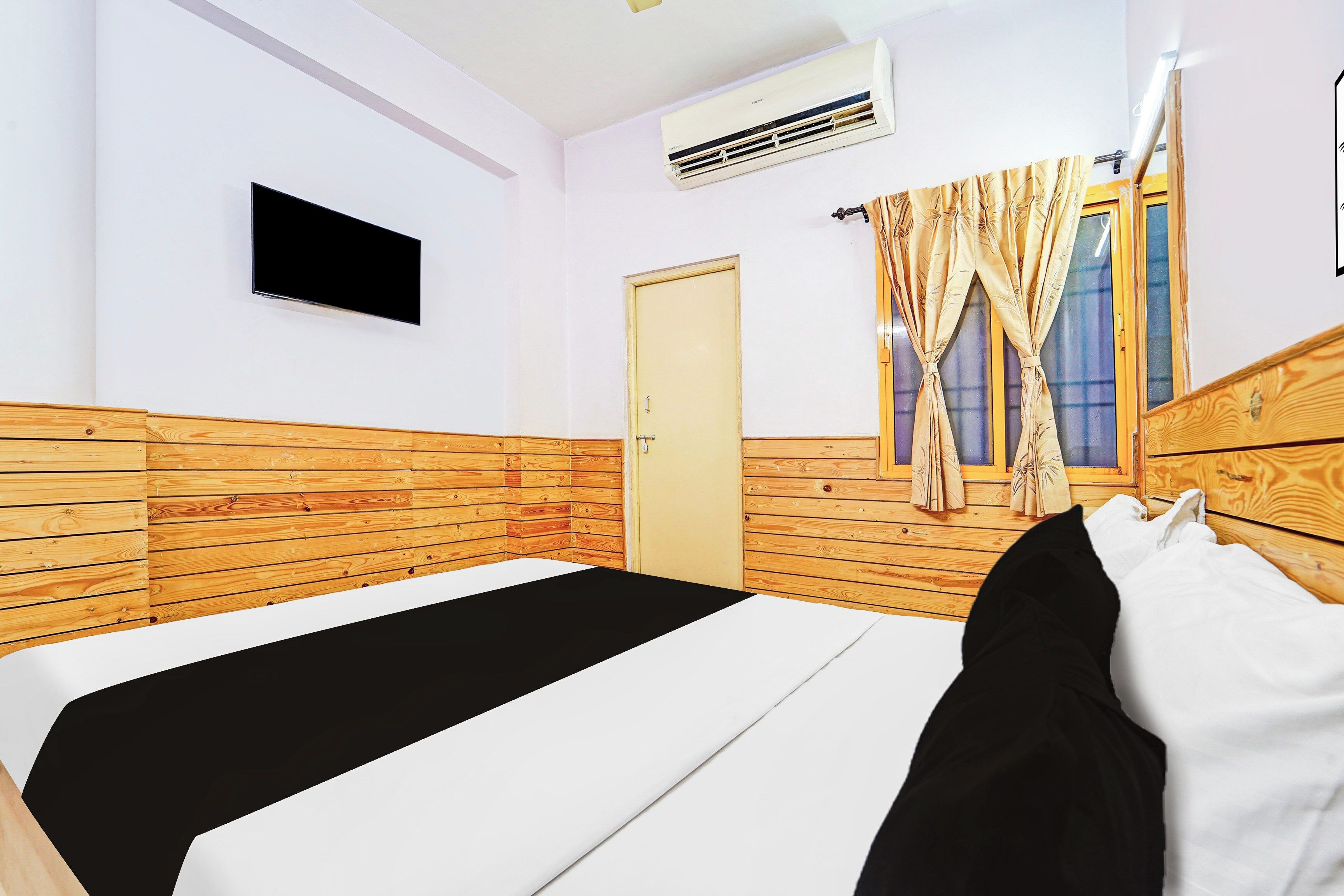 Een hotelkamer met een houten hoofdeinde, een enkel bed met een zwart-wit dekbedovertrek, een flatscreen televisie aan de muur bevestigd, en een raam met gordijnen.