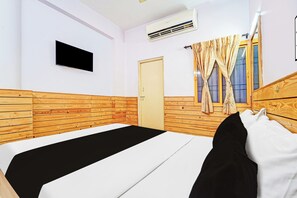 A hotel room with a wooden headboard, a single bed with a black and white bedspread, a flat-screen TV mounted on the wall, and a window with curtains.