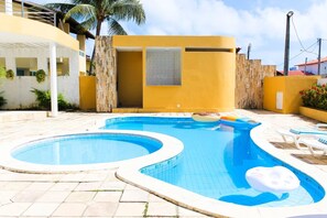 A swimming pool with two separate sections, surrounded by a yellow building with a modern design, and a palm tree in the background. - New Listing (Tamandaré)