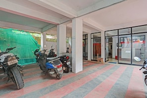 Un área de estacionamiento techada con varias scooters estacionadas, una zona delimitada con una red verde y la entrada de un edificio con puertas de vidrio.