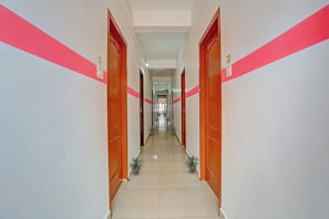 Un pasillo largo y estrecho con puertas a ambos lados, que conduce a habitaciones. El pasillo tiene suelo de baldosas y está bordeado de plantas en macetas.
