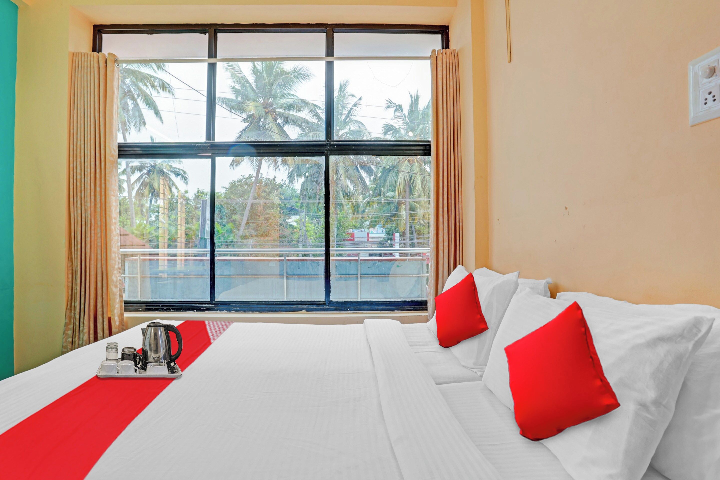 A hotel room with a large bed, red pillows, a coffee maker, and a view of palm trees through a window.