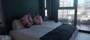 A hotel room with a bed, two decorative pillows, a window with a view of buildings, and a chair outside. - Luxurious 2-Bedroom Apartment in uMhlanga, South Africa (uMhlanga)