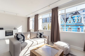 En moderne stue med sofa, sofabord og udsigt til en blå bygning gennem store vinduer.