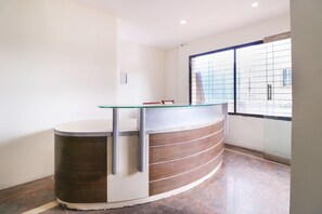 Balcão de recepção com tampo de vidro e painéis de madeira, uma cadeira e uma janela com vista para o exterior.