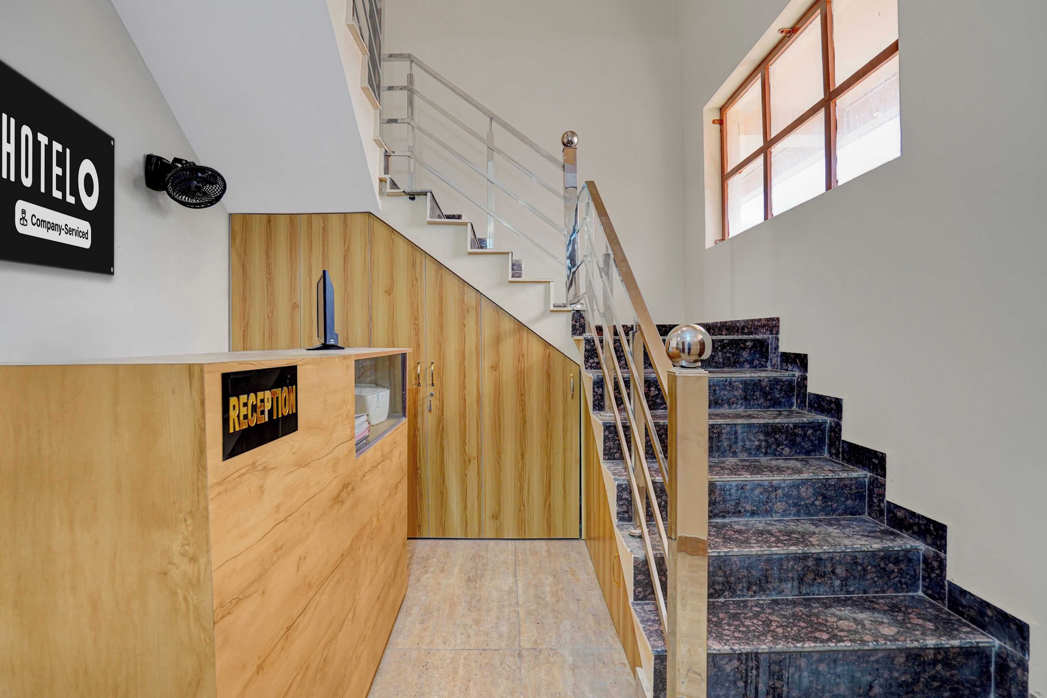 Una recepción con detalles de madera y un letrero que indica un hotel de servicio empresarial. Una escalera con barandilla de vidrio y pasamanos de madera conduce a un rellano con ventana.