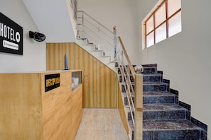 Un comptoir d’accueil avec un revêtement en bois et une enseigne indiquant qu’il s’agit d’un hôtel géré par une entreprise. Un escalier avec une rampe en verre et une main courante en bois mène à un palier doté d’une fenêtre.