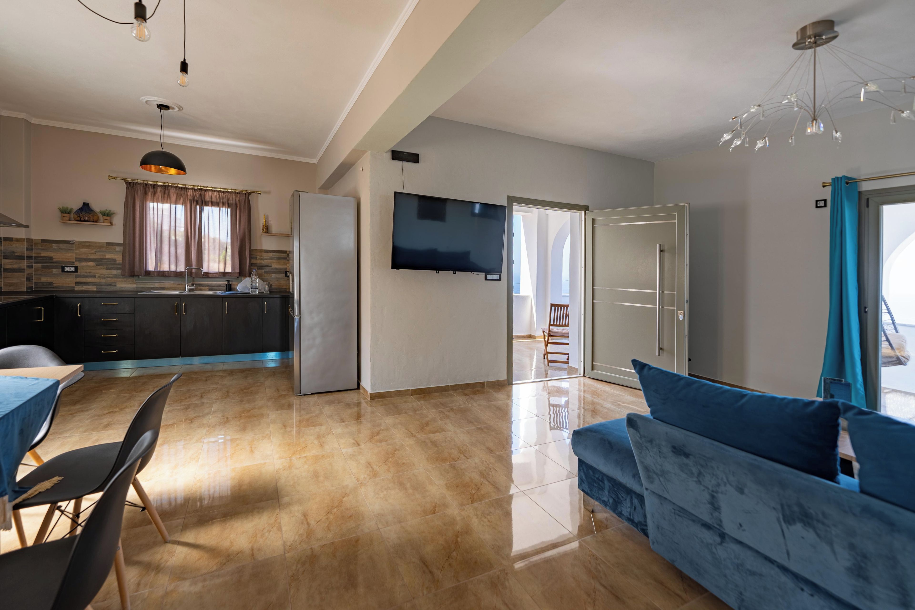 A modern kitchen with a refrigerator, a dining area with a table and chairs, a living room with a sofa, and a TV mounted on the wall.