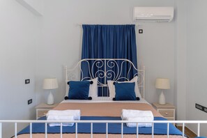 A bedroom with a bed, two bedside tables with lamps, a blue curtain, and an air conditioning unit. - Loulaki Villas Santorini by MPS (vourvoulos)