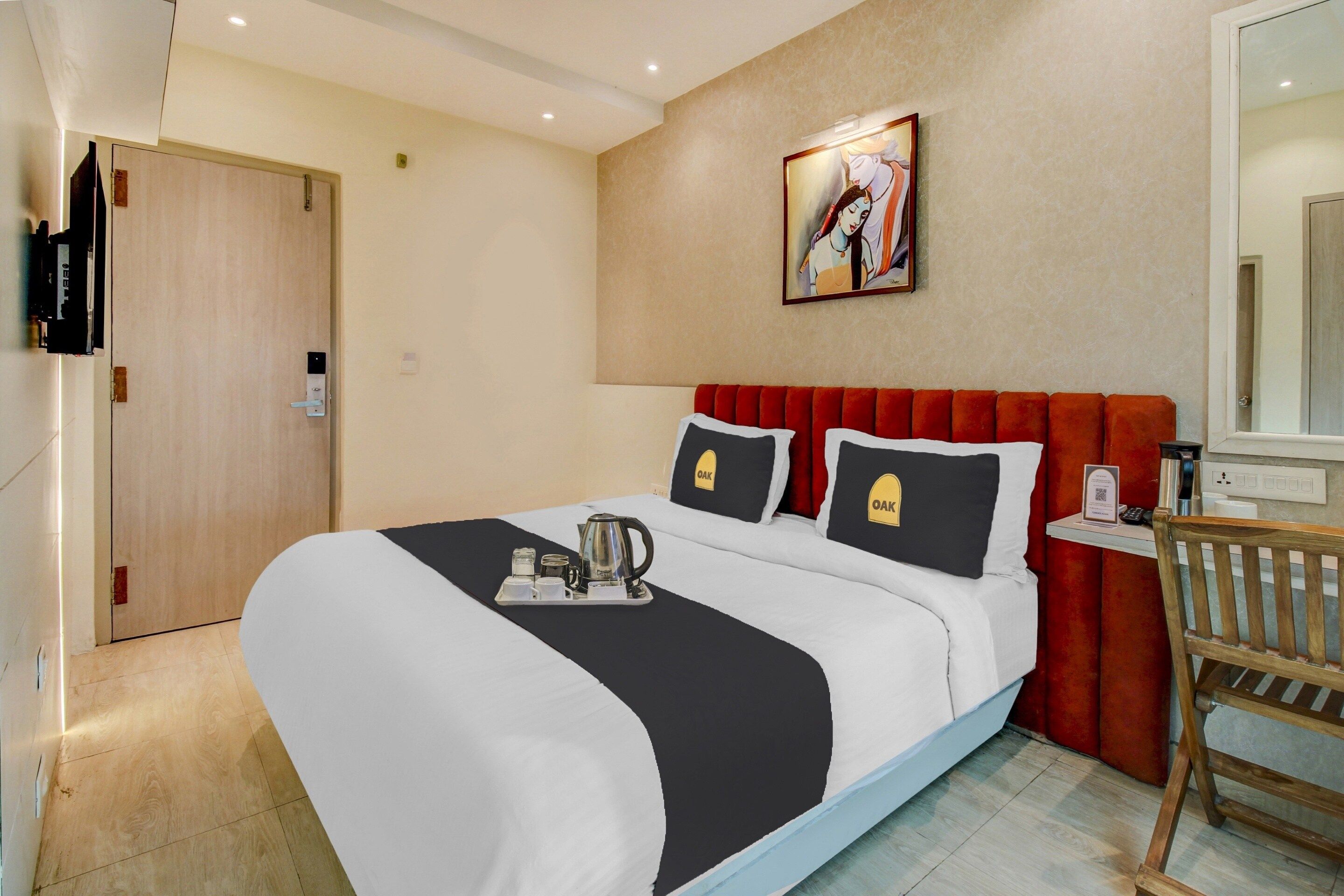 Habitación de hotel con una cama, un cuadro en la pared, una puerta de madera y una mesita con una tetera y tazas.