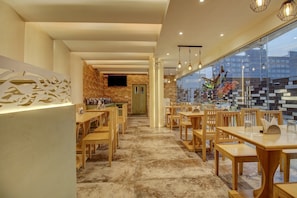 A restaurant interior with wooden tables and chairs, a brick wall, and a large window.