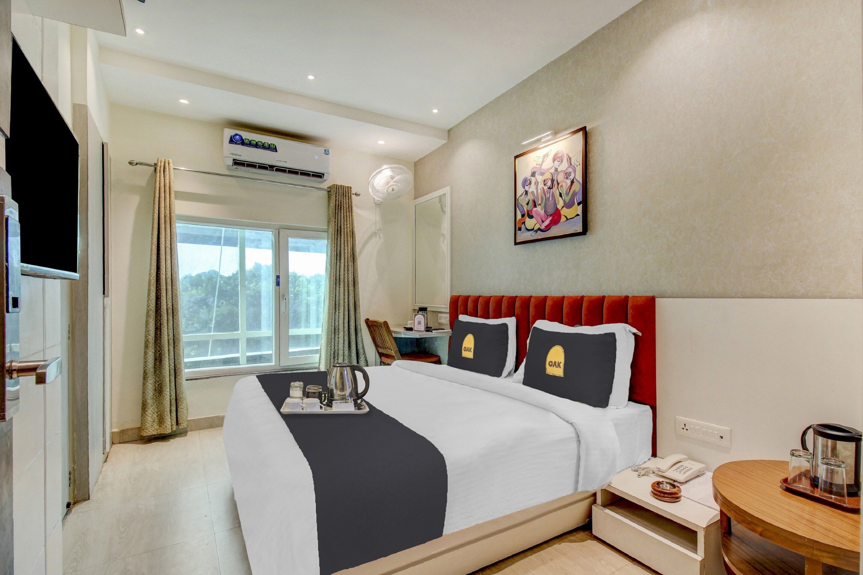 A hotel room with a bed, a bedside table with a phone and a kettle, a window with curtains, an air conditioning unit, and a painting on the wall.