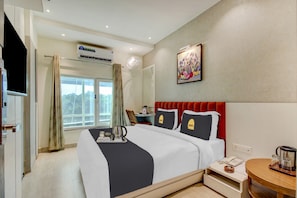 A hotel room with a bed, a bedside table with a phone and a kettle, a window with curtains, an air conditioning unit, and a painting on the wall.