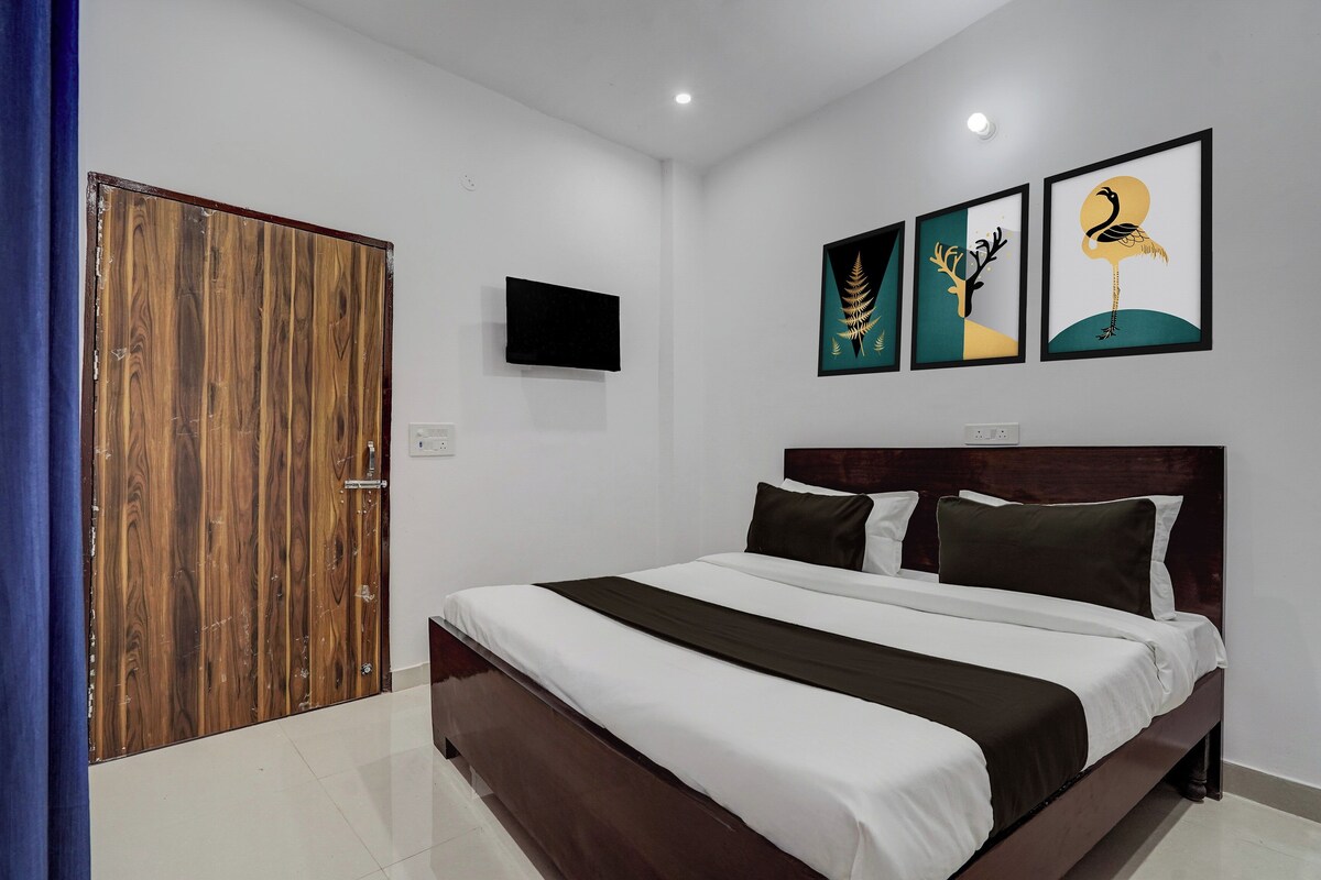 A bedroom with a wooden door, a bed with white and dark bedding, and three framed artworks on the wall.